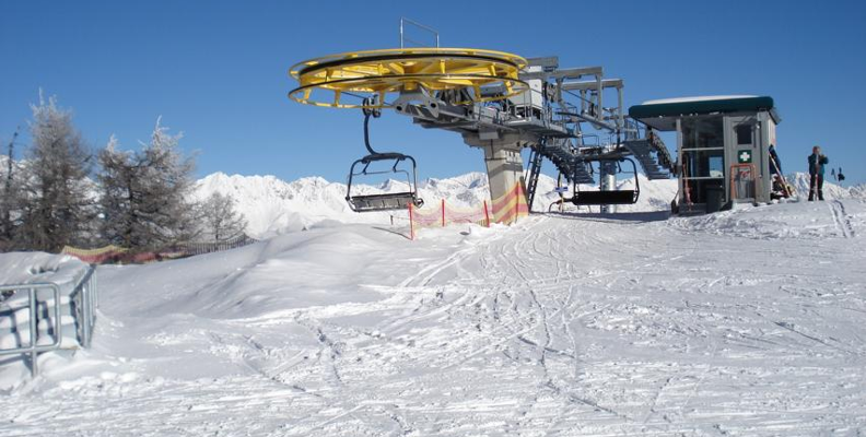Eine Skiliftstation auf einem verschneiten Berg mit leeren Sesseln, schneebedecktem Boden und entfernten Bergen unter einem klaren blauen Himmel.