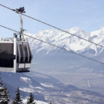 Eine Seilbahn fährt über eine verschneite Berglandschaft mit Kiefern und entfernten Tälern unter einem klaren Himmel.