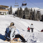 Menschen, die sich auf einer verschneiten Piste in der Nähe einer Holzhütte vergnügen, mit Sesselliften über dem Kopf und Bergen im Hintergrund.