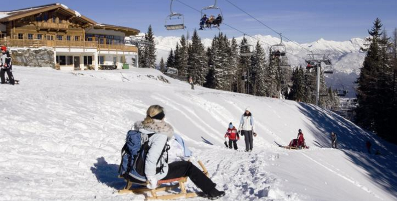 Menschen, die sich auf einer verschneiten Piste in der Nähe einer Holzhütte vergnügen, mit Sesselliften über dem Kopf und Bergen im Hintergrund.