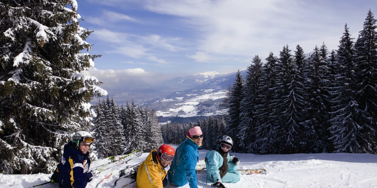 Vier Personen in Skiausrüstung sitzen auf einer verschneiten Piste, umgeben von Kiefern, mit einer Berglandschaft und einem teilweise bewölkten Himmel im Hintergrund.