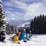 Vier Personen in Skiausrüstung sitzen auf einer verschneiten Piste, umgeben von Kiefern, mit einer Berglandschaft und einem teilweise bewölkten Himmel im Hintergrund.