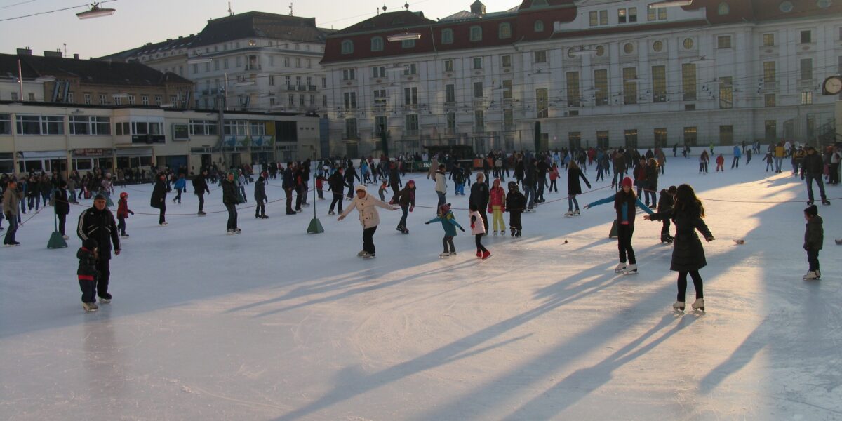 Viele Menschen laufen an einem sonnigen Tag auf einer Eisbahn im Freien vor großen Gebäuden Schlittschuh, wobei lange Schatten auf das Eis geworfen werden.