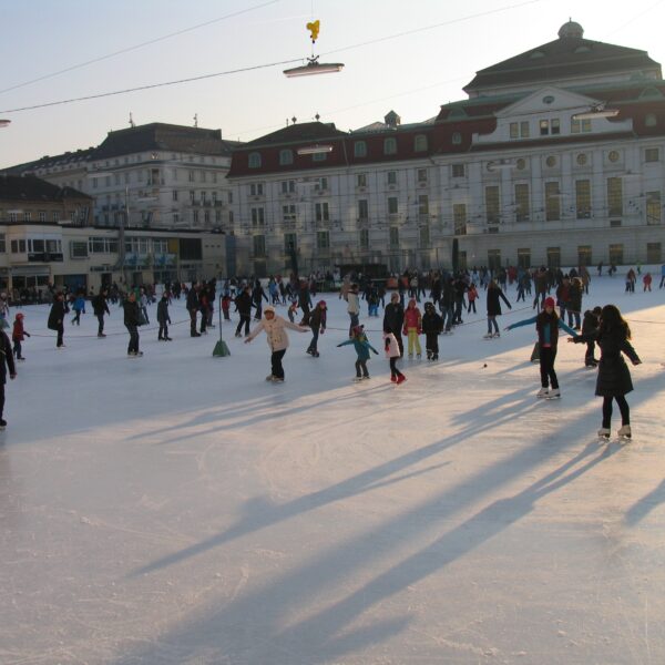 Viele Menschen laufen an einem sonnigen Tag auf einer Eisbahn im Freien vor großen Gebäuden Schlittschuh, wobei lange Schatten auf das Eis geworfen werden.
