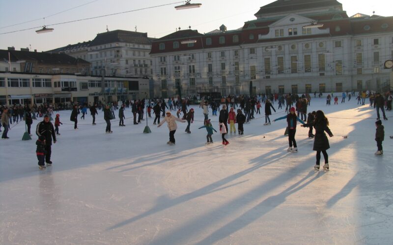 Viele Menschen laufen an einem sonnigen Tag auf einer Eisbahn im Freien vor großen Gebäuden Schlittschuh, wobei lange Schatten auf das Eis geworfen werden.