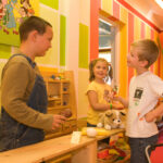 Drei Kinder spielen zusammen in einem farbenfrohen Raum; eines steht hinter einer Holztheke, während die beiden anderen Spielzeug halten und lächeln.