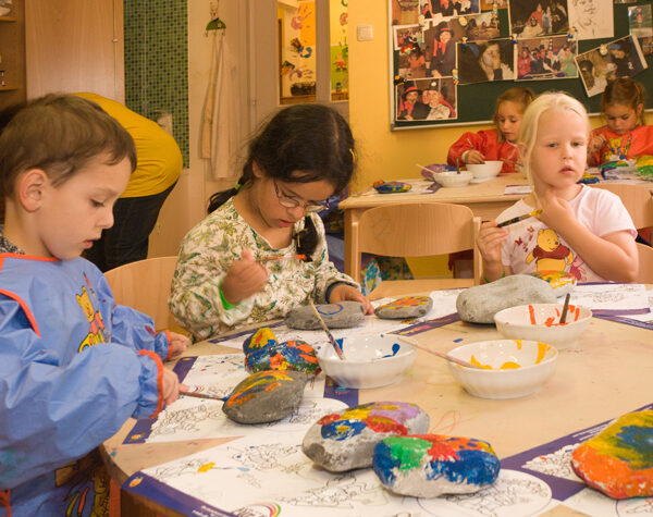 Drei kleine Kinder sitzen an einem Tisch in einem Klassenzimmer und bemalen große Felsen mit Pinseln und bunten Farben. Im Hintergrund sind andere Kinder und Kunstwerke zu sehen.