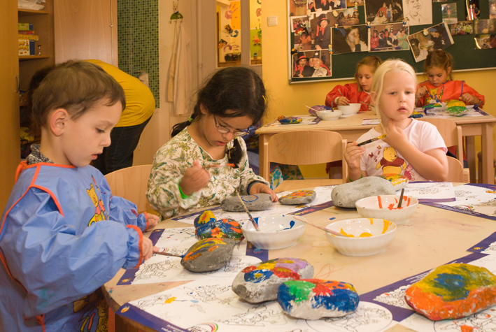 Drei kleine Kinder sitzen an einem Tisch in einem Klassenzimmer und bemalen große Felsen mit Pinseln und bunten Farben. Im Hintergrund sind andere Kinder und Kunstwerke zu sehen.