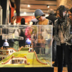 Drei Personen untersuchen ein architektonisches Miniaturmodell, das in einer Glasvitrine in einer Innenausstellung ausgestellt ist, während andere Besucher im Hintergrund zu sehen sind.