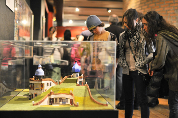 Drei Personen untersuchen ein architektonisches Miniaturmodell, das in einer Glasvitrine in einer Innenausstellung ausgestellt ist, während andere Besucher im Hintergrund zu sehen sind.