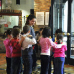 Eine Gruppe kleiner Kinder steht in einer Reihe in einem Gebäude, jedes Kind hat die Hände auf den Schultern des Kindes vor ihm, angeführt von einer erwachsenen Frau.