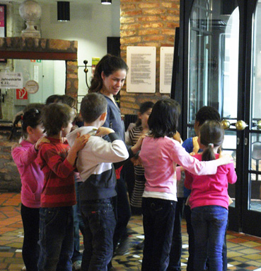 Eine Gruppe kleiner Kinder steht in einer Reihe in einem Gebäude, jedes Kind hat die Hände auf den Schultern des Kindes vor ihm, angeführt von einer erwachsenen Frau.