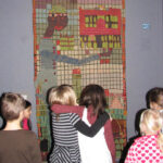 Eine Gruppe von Kindern steht vor einem farbenfrohen, abstrakten Wandteppich, auf dem eine hochgewachsene Figur mit einem verlängerten Arm abgebildet ist und der an einer Wand hängt.