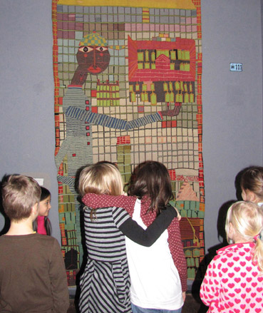 Eine Gruppe von Kindern steht vor einem farbenfrohen, abstrakten Wandteppich, auf dem eine hochgewachsene Figur mit einem verlängerten Arm abgebildet ist und der an einer Wand hängt.