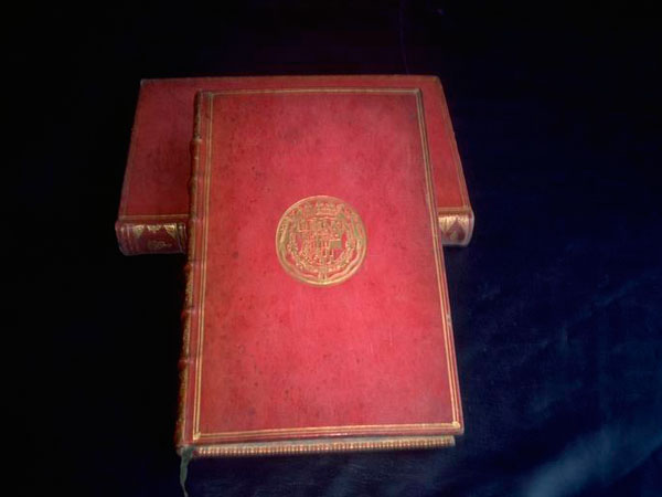 Zwei alte, in rotes Leder gebundene Bücher mit goldenen Verzierungen und einem geprägten Wappen auf dem vorderen Einband, die auf einer dunklen Fläche stehen.