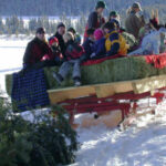 Eine Gruppe von Menschen, darunter Kinder, fährt auf einem von Pferden gezogenen Heuwagen durch den Schnee, umgeben von Bäumen in einer Winterlandschaft.