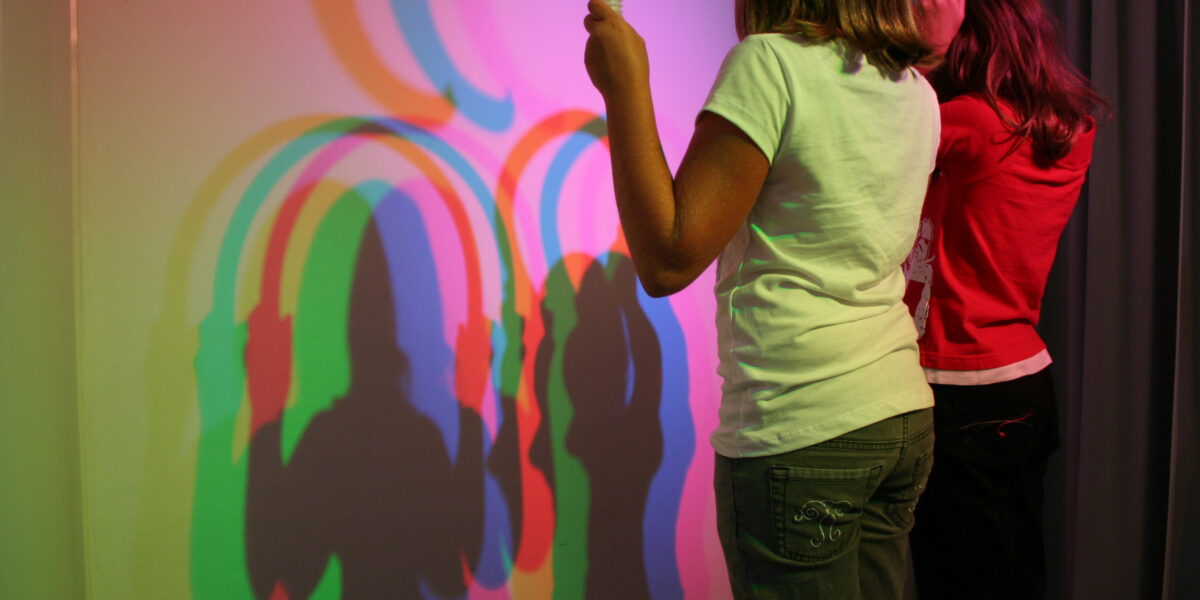 Zwei Kinder halten Röhren vor ein Licht und erzeugen dadurch überlappende bunte Schatten an einer Wand hinter ihnen.