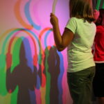 Zwei Kinder halten Röhren vor ein Licht und erzeugen dadurch überlappende bunte Schatten an einer Wand hinter ihnen.