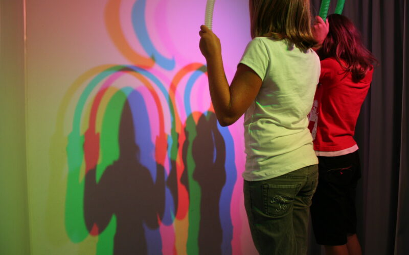 Zwei Kinder halten Röhren vor ein Licht und erzeugen dadurch überlappende bunte Schatten an einer Wand hinter ihnen.