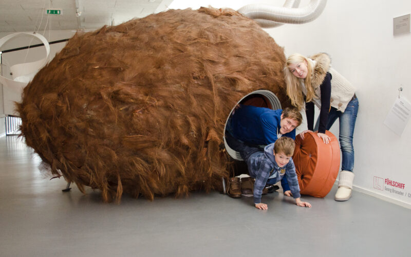 Drei Personen interagieren im Innenbereich mit einer großen, pelzigen, braunen, tunnelartigen Skulptur; zwei Kinder krabbeln heraus, während ein Erwachsener am Eingang lehnt.