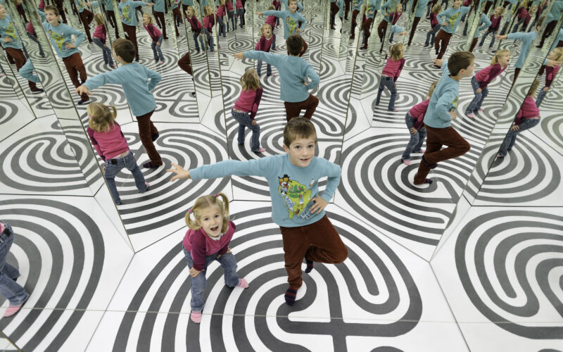 Zwei Kinder stehen auf einem schwarz-weiß gemusterten Boden, umgeben von verspiegelten Wänden, und erzeugen mehrere Spiegelbilder von sich selbst in verschiedenen Posen.