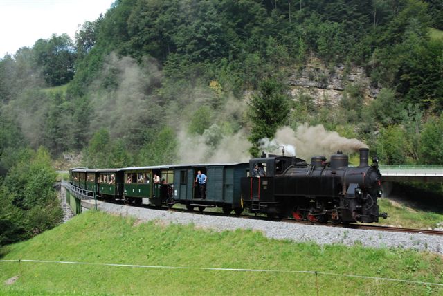 Ein alter Dampfzug mit mehreren grünen Waggons fährt durch eine üppige, hügelige Landschaft und stößt Rauchwolken aus.