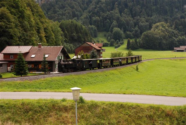 Ein Oldtimerzug fährt an Häusern vorbei und durch grüne Landschaften mit Bäumen und Hügeln im Hintergrund.