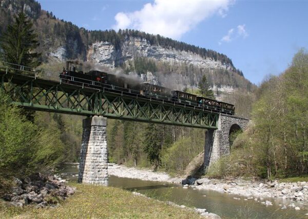 Ein schwarzer Dampfzug überquert eine grüne Metallbrücke über einen Fluss, mit felsigen Klippen und Bäumen im Hintergrund unter einem teilweise bewölkten Himmel.