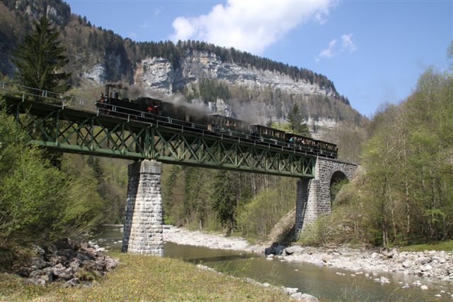Ein schwarzer Dampfzug überquert eine grüne Metallbrücke über einen Fluss, mit felsigen Klippen und Bäumen im Hintergrund unter einem teilweise bewölkten Himmel.