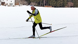 Ein älterer Mann in gelber Jacke und schwarzer Hose beim Skilanglauf auf einem verschneiten Feld mit Bäumen und Gebäuden im Hintergrund.