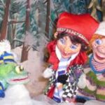 Drei Puppen - ein Frosch mit blauem und gelbem Schal und zwei Kinder in Winterkleidung - stehen vor einer verschneiten Waldkulisse.