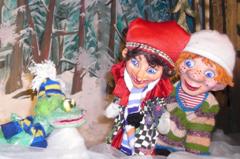Drei Puppen - ein Frosch mit blauem und gelbem Schal und zwei Kinder in Winterkleidung - stehen vor einer verschneiten Waldkulisse.