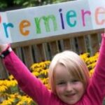 Ein kleines Kind in einem rosa Hemd hält ein buntes Schild mit der Aufschrift "Premiere" vor gelben Blumen und einem Holzzaun hoch.