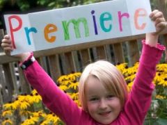 Ein kleines Kind in einem rosa Hemd hält ein buntes Schild mit der Aufschrift "Premiere" vor gelben Blumen und einem Holzzaun hoch.