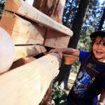 Ein kleines Kind in blauem Hemd und Mütze steht lächelnd an einem Holzgerüst im Freien, im Hintergrund sind Bäume zu sehen.