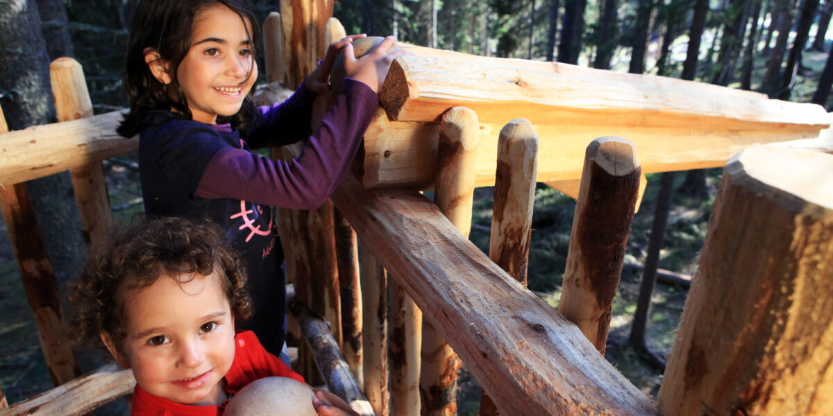 Zwei Kinder stehen in einer Holzkonstruktion in einem Wald; eines hält einen Ball und schaut in die Kamera, während das andere sich lächelnd am Geländer festhält.