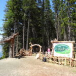 Ein hölzernes Eingangstor und ein Schild mit einer Landkarte stehen am Rande eines Waldes; zwei Personen stehen in der Nähe des Eingangs, im Hintergrund sind Berge zu sehen.