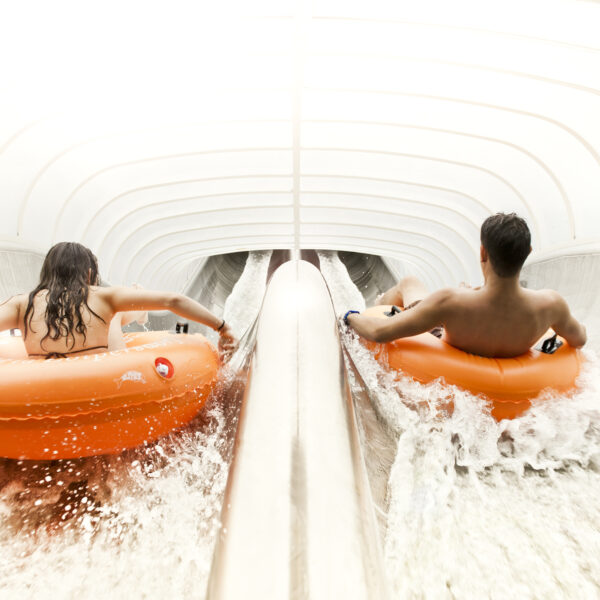 Zwei Personen fahren mit orangefarbenen Schläuchen auf parallelen Wasserrutschen in einem hell beleuchteten, geschlossenen Wasserpark.