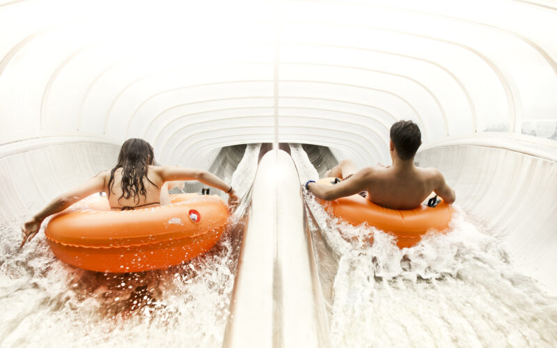 Zwei Personen fahren mit orangefarbenen Schläuchen auf parallelen Wasserrutschen in einem hell beleuchteten, geschlossenen Wasserpark.