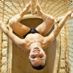 Ein lächelnder Junge hängt kopfüber von einer Seilbrücke und hält sich mit Beinen und Armen an den Seilen fest.