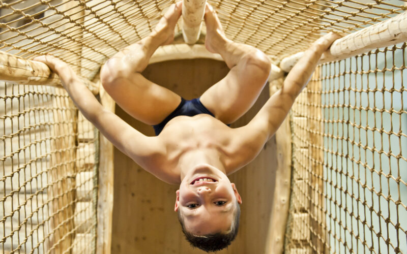 Ein lächelnder Junge hängt kopfüber von einer Seilbrücke und hält sich mit Beinen und Armen an den Seilen fest.