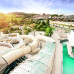 Luftaufnahme eines Wasserparks mit mehreren großen Röhrenrutschen, einem Schwimmbecken und einer umgebenden grünen Landschaft mit Gebäuden im Hintergrund.