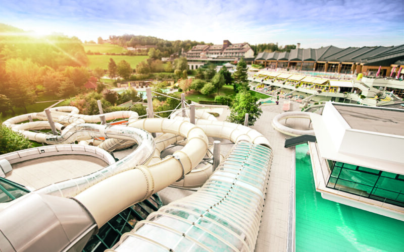 Luftaufnahme eines Wasserparks mit mehreren großen Röhrenrutschen, einem Schwimmbecken und einer umgebenden grünen Landschaft mit Gebäuden im Hintergrund.