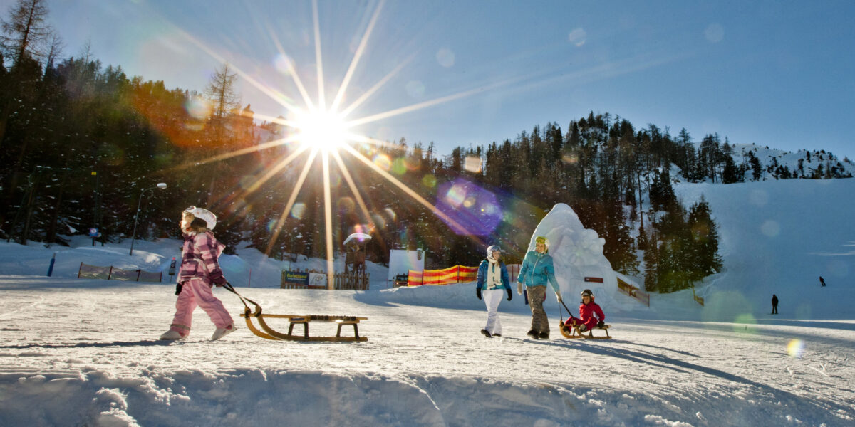 Kinder in Winterkleidung laufen und spielen mit Schlitten auf einem verschneiten Hang unter der strahlenden Sonne, umgeben von Bäumen und fernen Bergen.