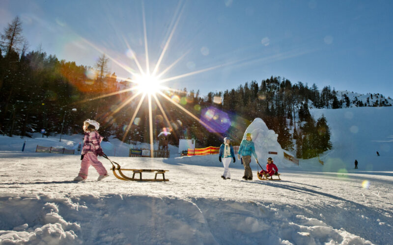 Kinder in Winterkleidung laufen und spielen mit Schlitten auf einem verschneiten Hang unter der strahlenden Sonne, umgeben von Bäumen und fernen Bergen.