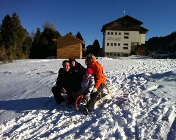 Vier Personen sitzen auf einem Holzschlitten im Schnee in der Nähe eines Gebäudes, mit Bäumen und einem klaren blauen Himmel im Hintergrund.
