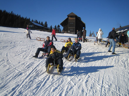 Menschen, die an einem sonnigen Tag einen verschneiten Hügel hinunterrodeln, mit einem Holzgebäude und mehreren anderen im Hintergrund.