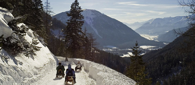 Vier Personen fahren auf Schlitten einen schneebedeckten, von Bäumen gesäumten Bergweg hinunter, mit Blick auf ferne Berge und ein Tal.