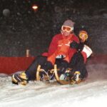 Zwei Personen in Winterkleidung und mit Schutzbrille fahren nachts auf einem Holzschlitten einen verschneiten Hang hinunter.
