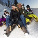Eine vierköpfige Familie in Winterkleidung und mit Helmen fährt in der Abenddämmerung auf Holzschlitten einen verschneiten Hügel hinunter.